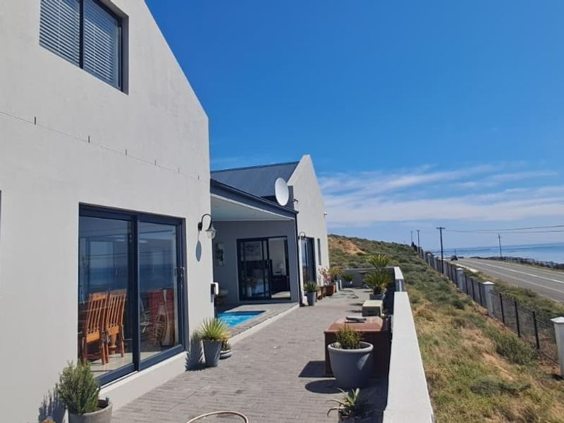 Rear exterior view of the property featuring a paved patio area, sliding glass doors, and a boundary wall overlooking the coastal road and ocean.