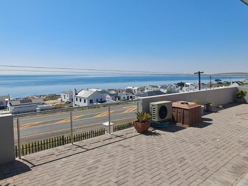 Spacious elevated balcony featuring paved flooring, a modern glass balustrade, and expansive coastal views overlooking the neighborhood and ocean.
