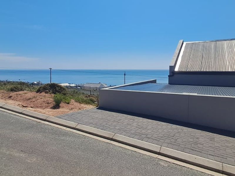 Front exterior view of the property featuring a modern corrugated metal roof, paved driveway, and boundary wall with a scenic coastal outlook.