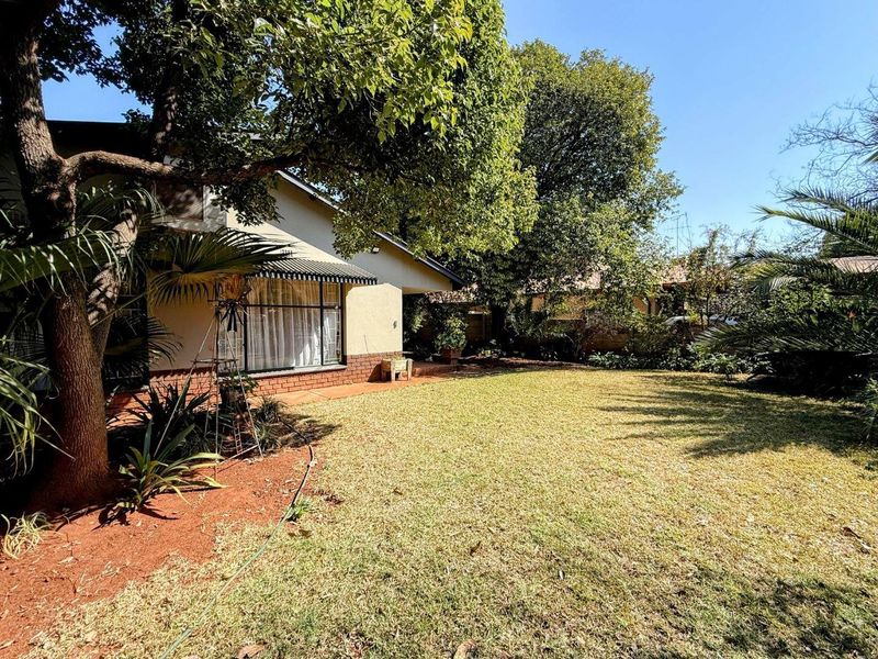 A view of the rear garden featuring a paved patio area, mature trees, and a well-maintained lawn adjacent to the house facade.