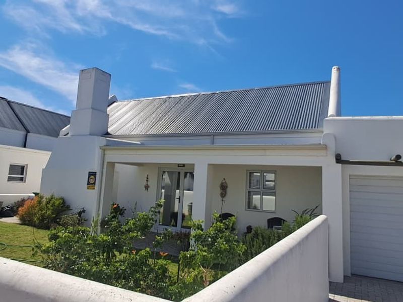 Front elevation of a well-maintained house featuring a corrugated metal roof, covered patio, attached garage, and a neat front garden with boundary wall.