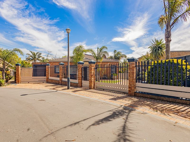 Front view of the property featuring a secure brick and metal perimeter fence, paved driveway access, and established landscaping.