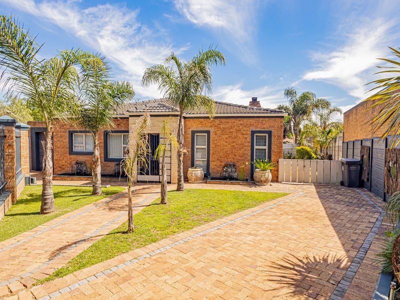 A well-maintained single-story brick house featuring a paved driveway, landscaped front garden, and secure gated entrance.