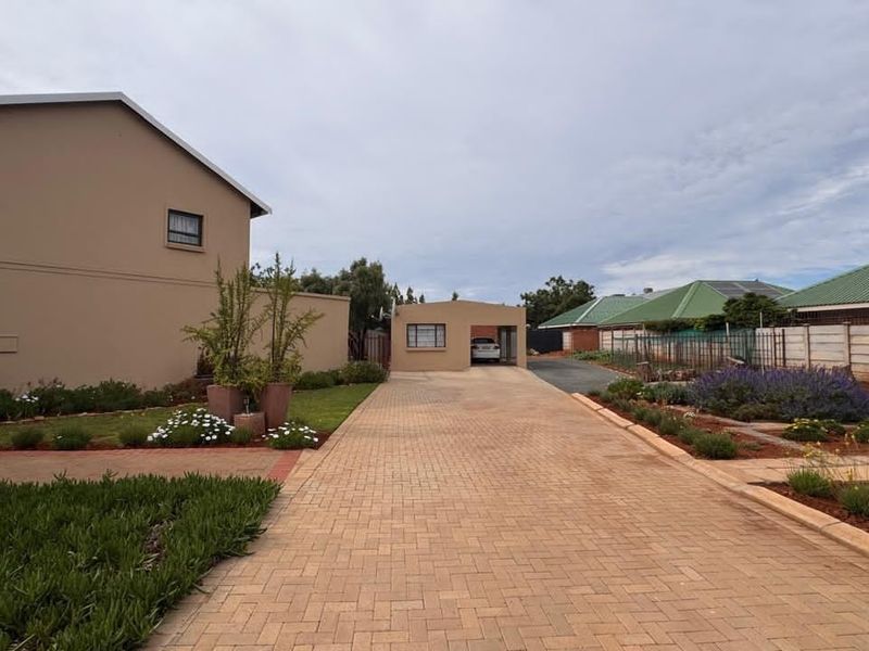 A well-maintained front exterior view featuring a long paved driveway, a carport, and neatly landscaped garden beds.