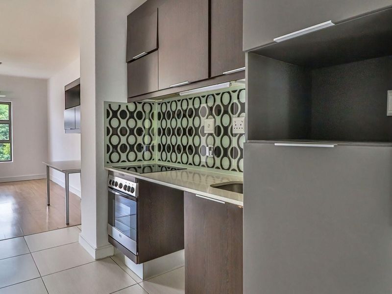 A modern kitchen featuring dark wood cabinetry, a patterned glass splashback, and integrated oven and hob, flowing into an open-plan living area.
