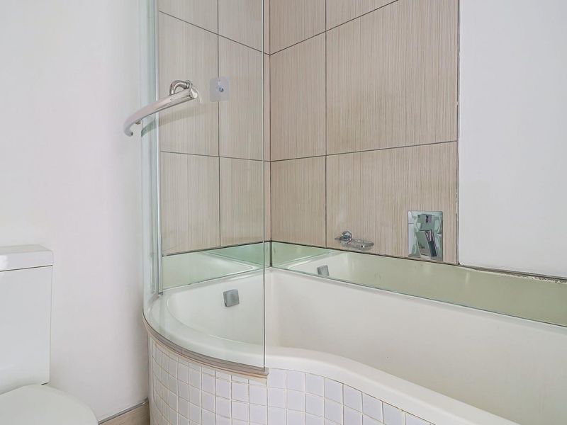 A functional bathroom featuring a curved bathtub with a glass shower screen and neutral ceramic wall tiling.