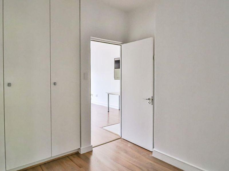 A bright, empty bedroom featuring built-in cupboards and light-toned laminate flooring, with a doorway leading to an adjacent living area.