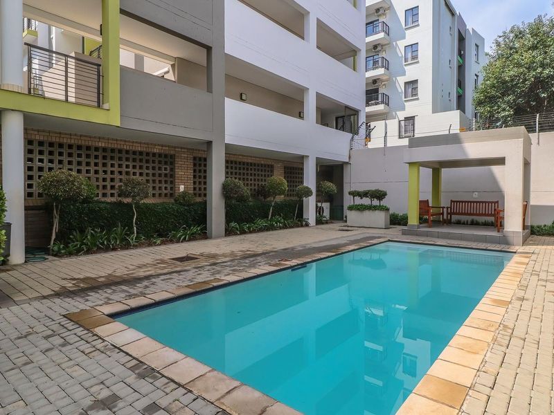 Communal swimming pool area featuring a paved deck, covered gazebo, and secure perimeter walling within the apartment complex.