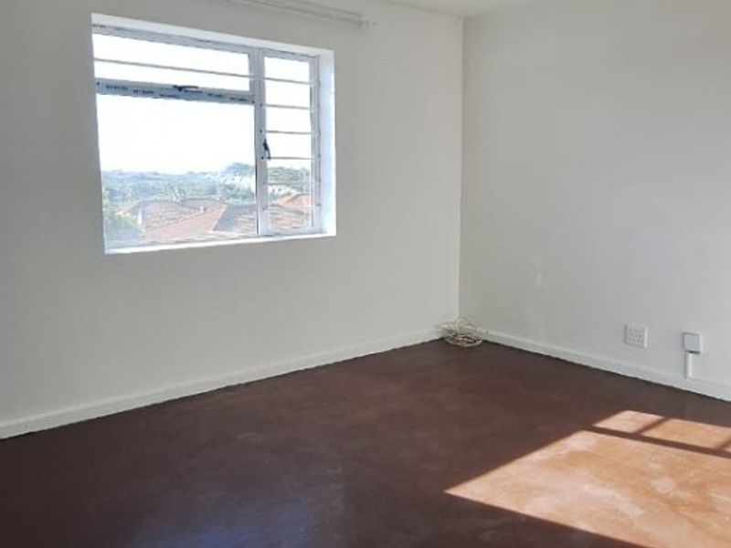 An empty bedroom featuring white walls, a single ceiling light, and an aluminium window frame with a view of the surrounding area.