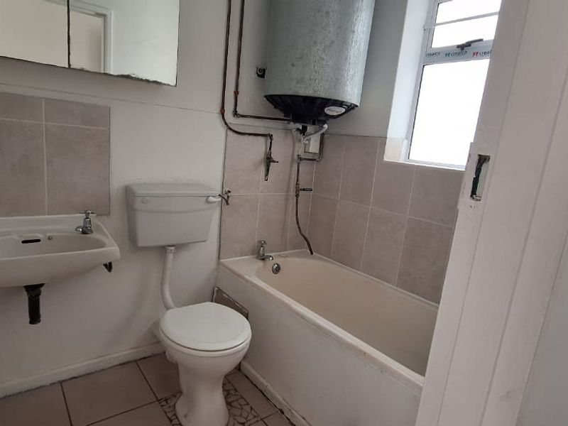 A functional bathroom featuring a bathtub, toilet, and wall-mounted basin, with exposed geyser plumbing and neutral tiled finishes.