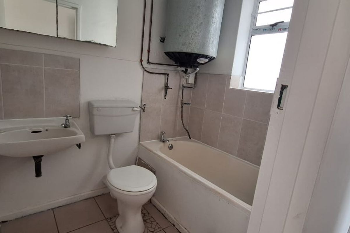 A functional bathroom featuring a bathtub, toilet, and wall-mounted basin, with exposed geyser plumbing and neutral tiled finishes.