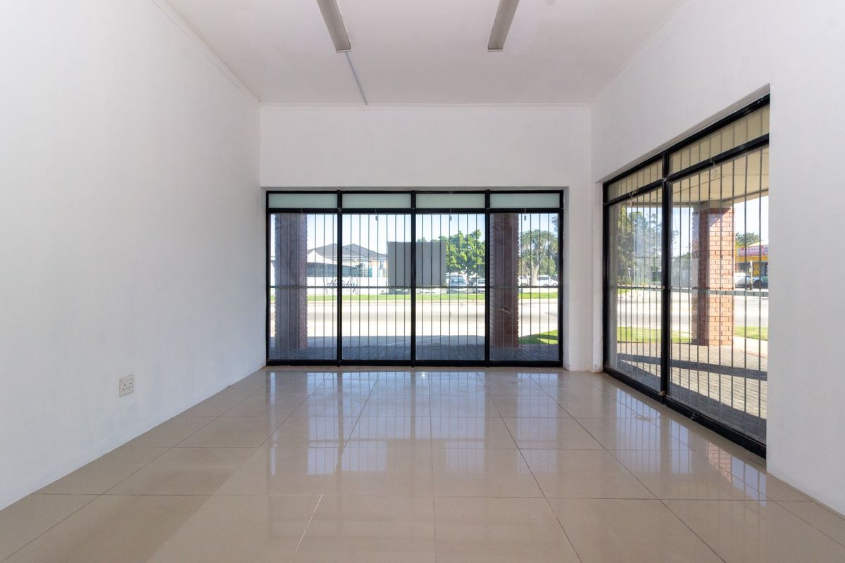 A bright, empty retail space featuring large glass windows with security bars, tiled flooring, and overhead fluorescent lighting, suitable for various commercial uses.