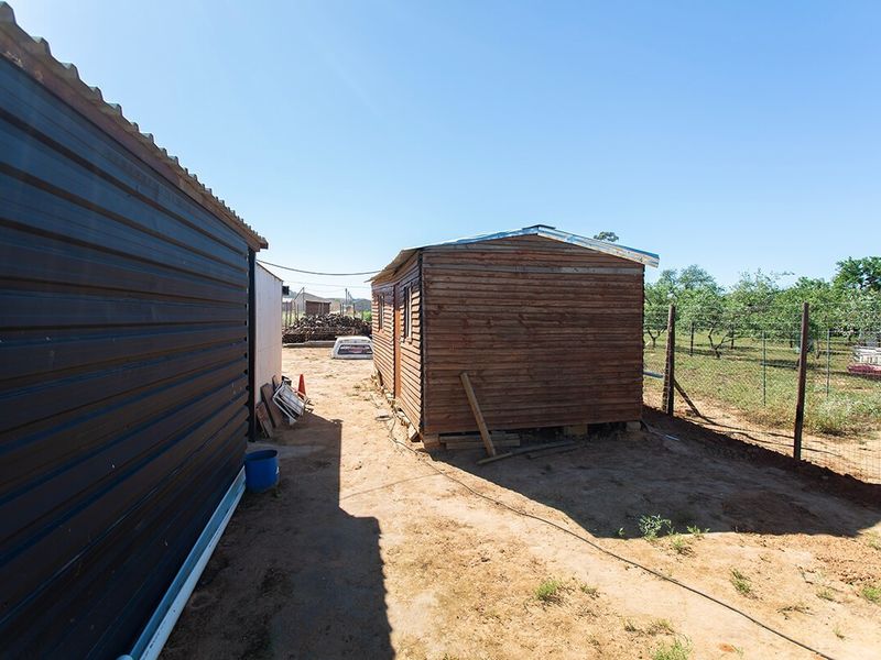 Rear exterior view of the small holding showing a corrugated metal structure and a timber shed set on a dirt yard with perimeter fencing.