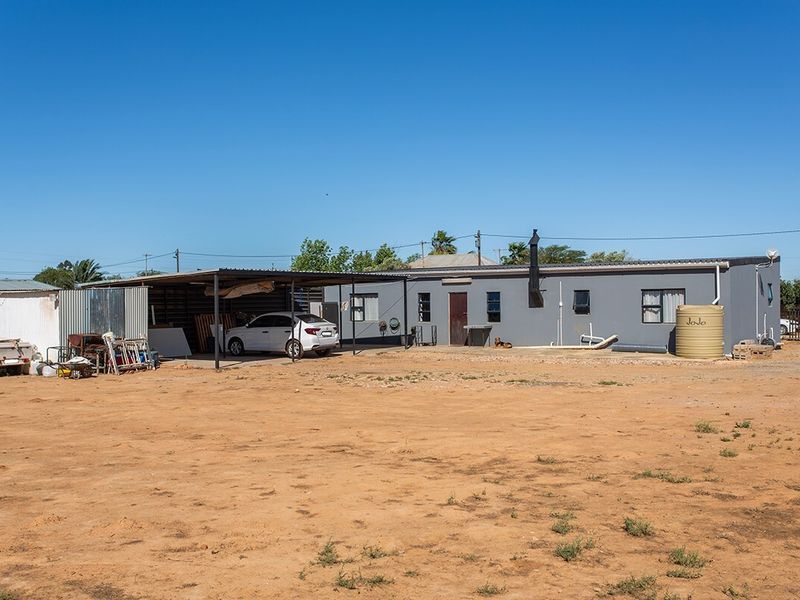 Rear view of the property featuring a large carport, exterior wall with flue pipe, and a water storage tank on a gravel and dirt yard.