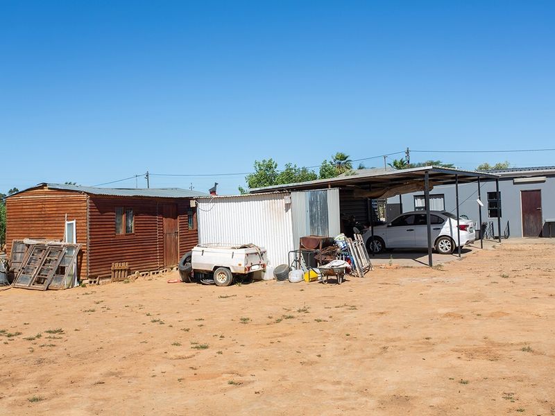 Rear view of the small holding featuring a timber-clad structure, a corrugated metal shed, and a covered carport area on an unpaved lot.