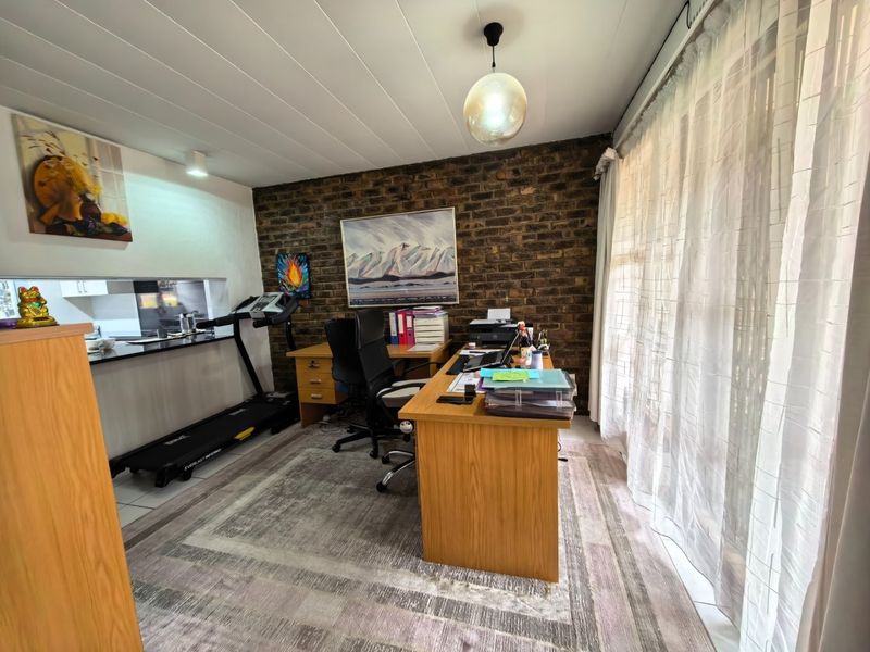 A functional home office space featuring an exposed brick feature wall, tiled flooring, and a convenient pass-through window connecting to the kitchen area.