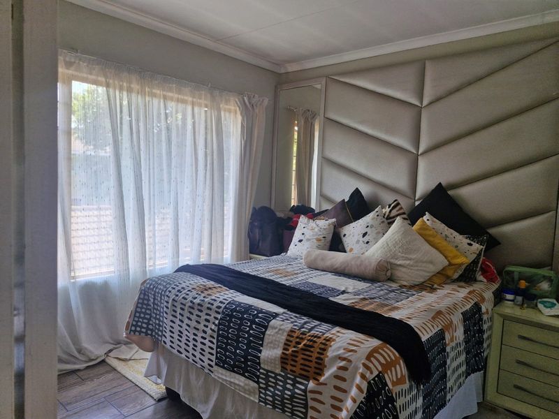 A comfortable bedroom featuring a large upholstered feature headboard wall, tiled flooring, and natural light through a large window.
