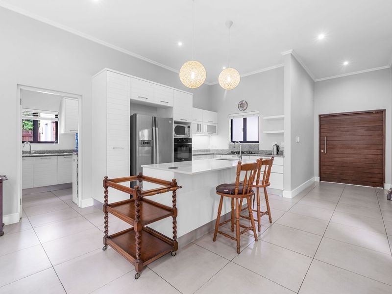 Modern open-plan kitchen featuring a central island, stone countertops, built-in appliances, and tiled flooring, with seamless access to an adjacent scullery area.