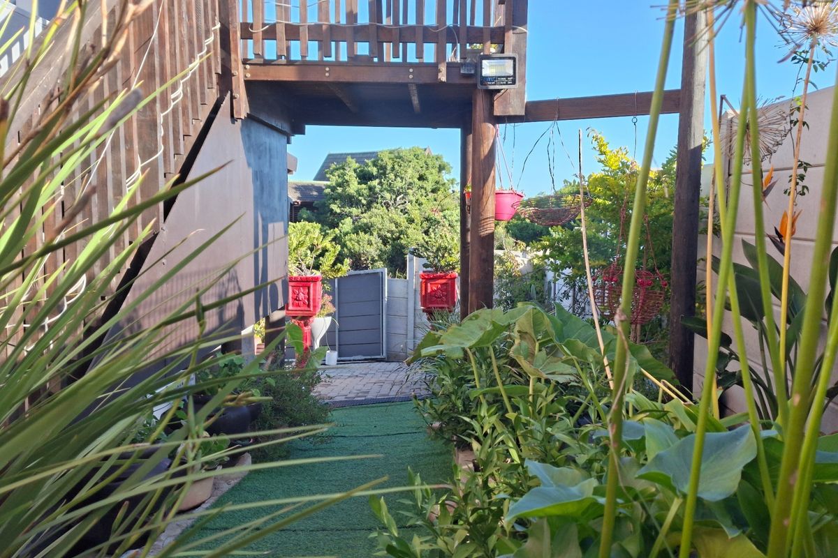 A rear garden area featuring a wooden staircase leading to an elevated deck, paved pathway, and established garden beds.