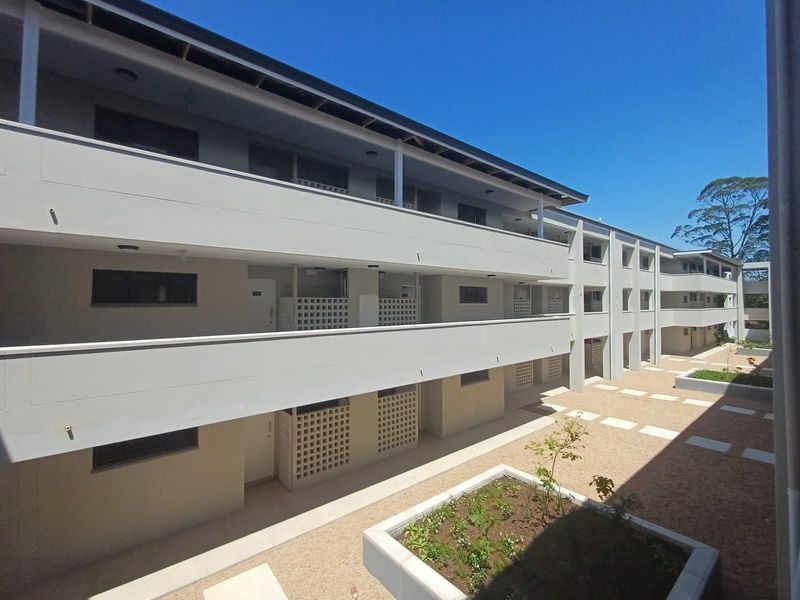 A modern multi-level apartment complex exterior featuring clean lines, concrete walkways, breeze block detailing, and a landscaped gravel courtyard area.