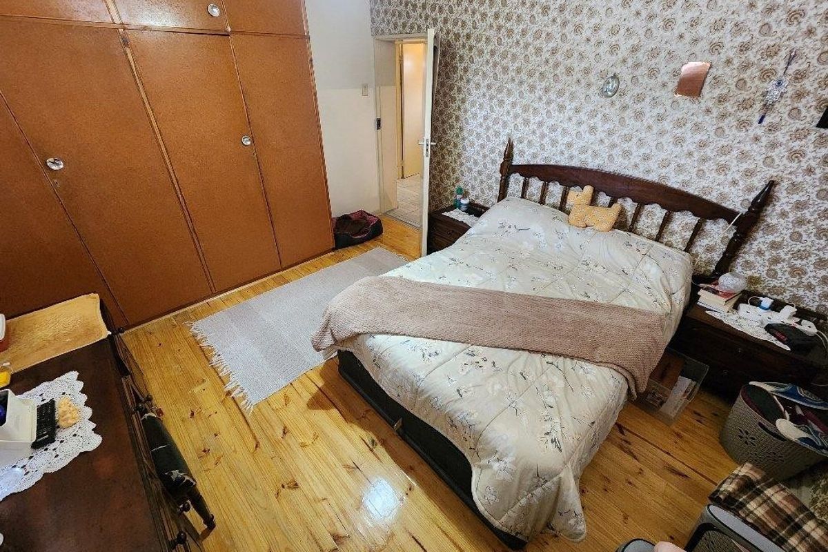 A bedroom featuring timber flooring, built-in cupboards, and dated patterned wallpaper, offering a functional space with potential for modernization.