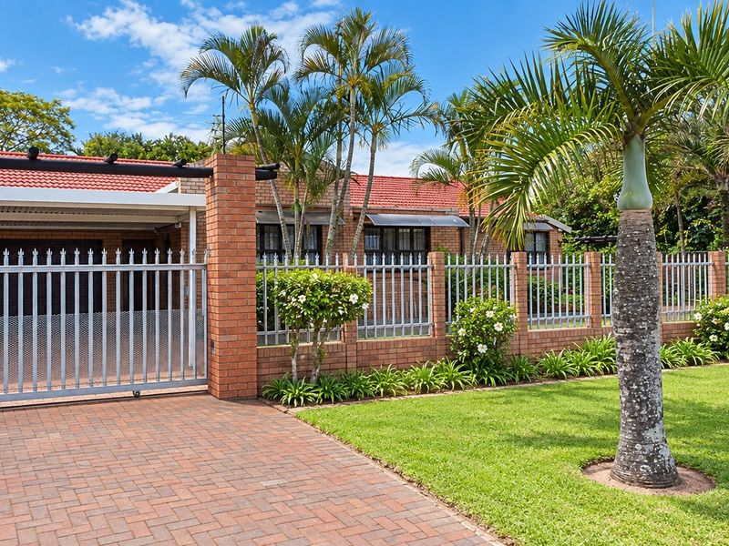 Front view of the property featuring a paved driveway, a covered carport, and a secure brick and metal perimeter fence with landscaped garden.