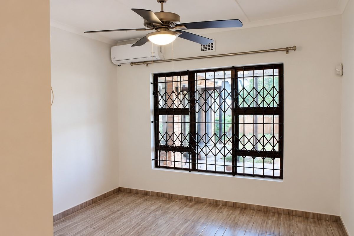 A clean, empty bedroom featuring tiled flooring, a ceiling fan, and a wall-mounted air conditioning unit with security bars on the window.
