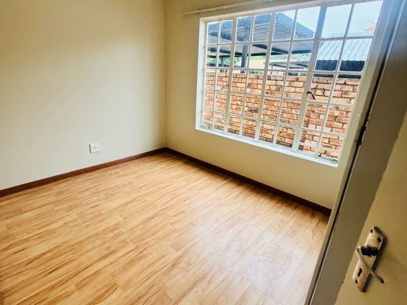 A bright, empty bedroom featuring light wood-look laminate flooring, neutral walls, and a large grid-pattern window providing natural light.