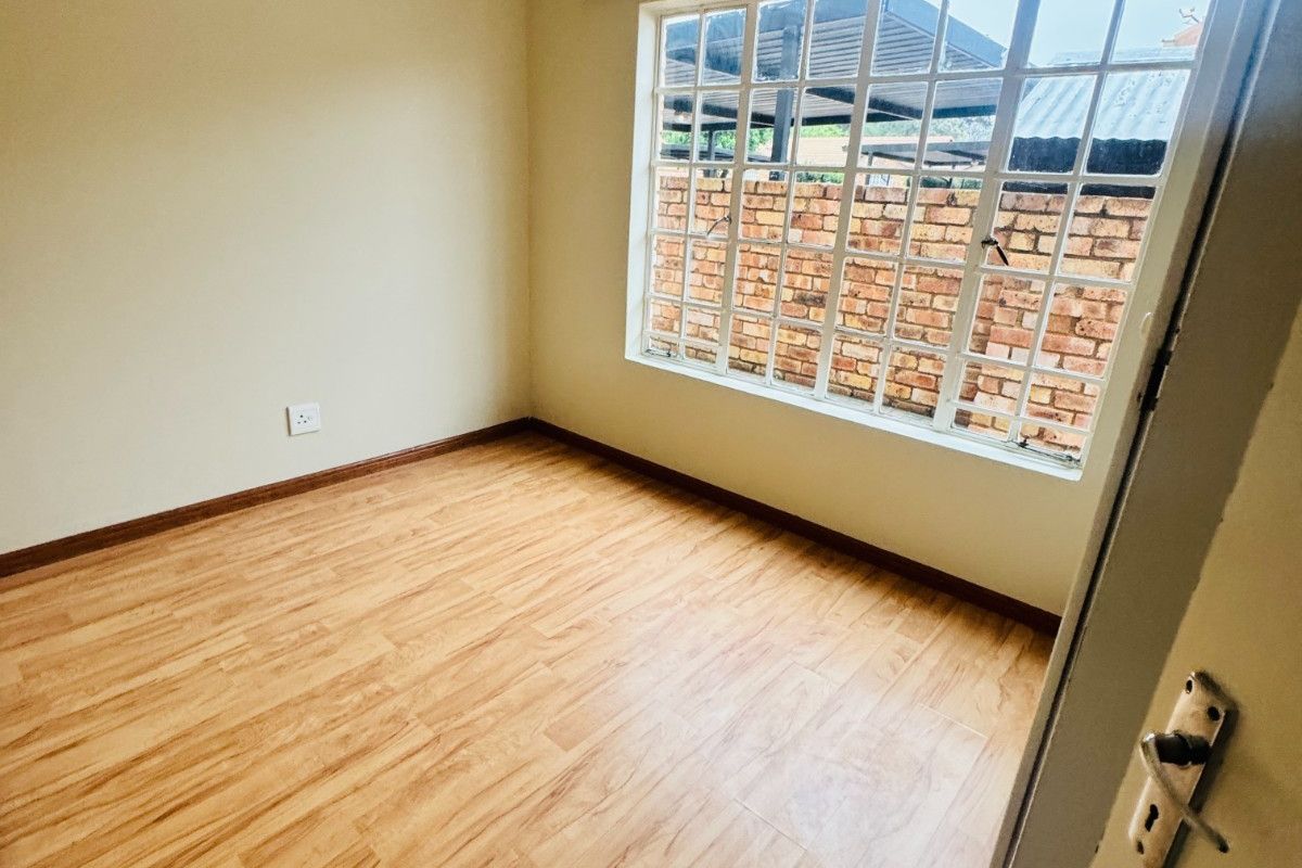 A bright, empty bedroom featuring light wood-look laminate flooring, neutral walls, and a large grid-pattern window providing natural light.