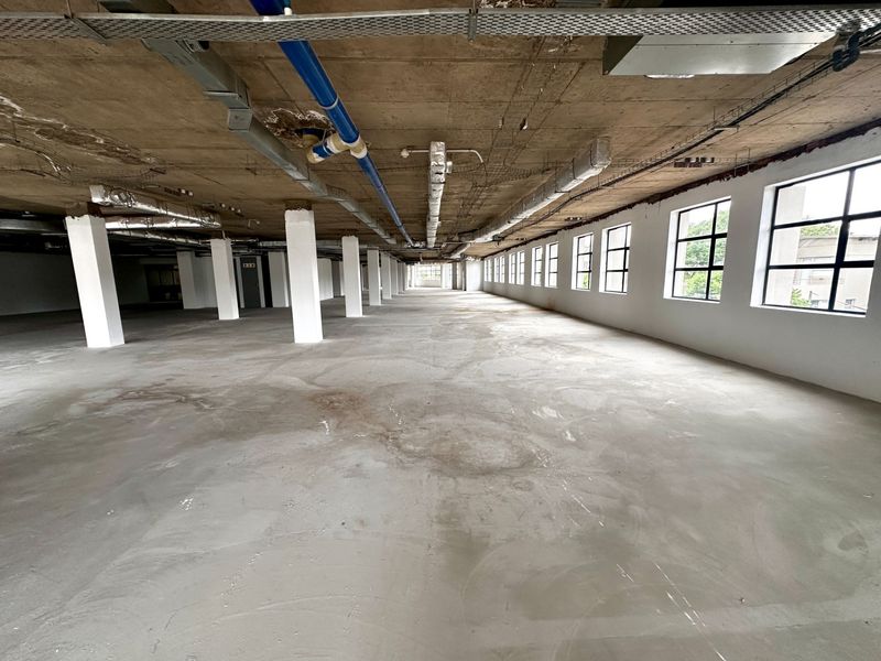 Expansive open-plan office space featuring an industrial aesthetic with exposed concrete ceilings, ductwork, and large windows providing ample natural light.