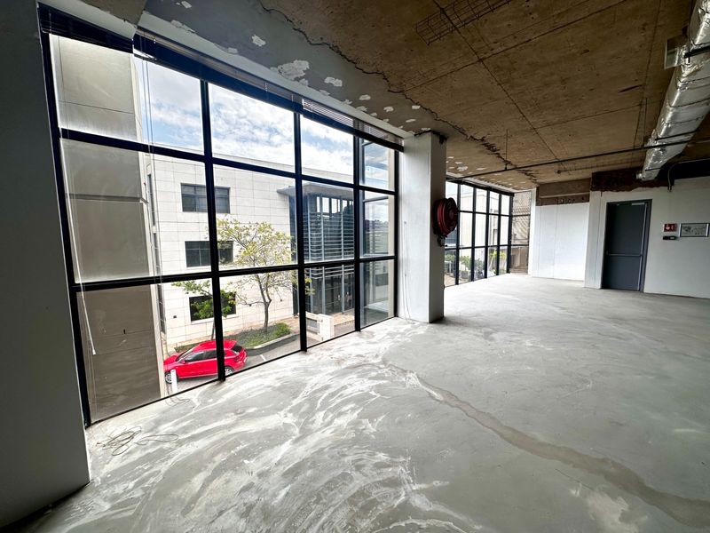An open-plan office space in shell condition featuring large floor-to-ceiling windows, exposed concrete ceilings, and industrial ducting, offering a blank canvas for customization.