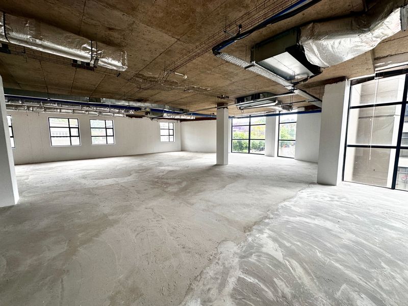 A spacious, open-plan office shell featuring exposed concrete ceilings, industrial HVAC ducting, and large windows providing ample natural light.
