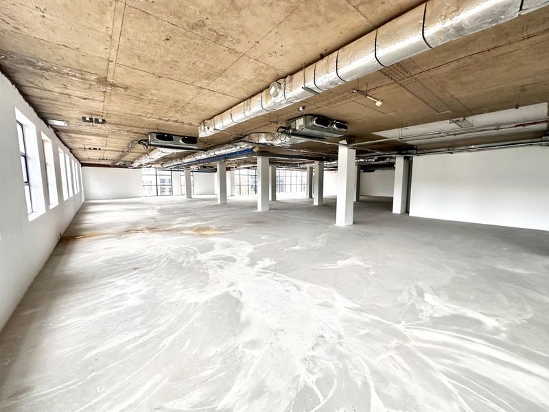 A large, open-plan office space featuring an industrial aesthetic with exposed concrete ceilings, visible HVAC ducting, and large steel-framed windows.