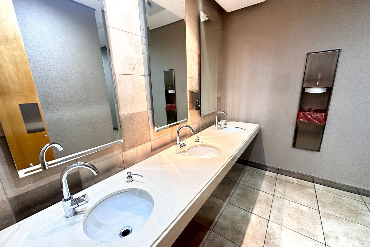 A clean, modern office bathroom featuring a triple vanity with undermount sinks, chrome fixtures, tiled backsplash, and integrated wall-mounted paper towel dispenser.