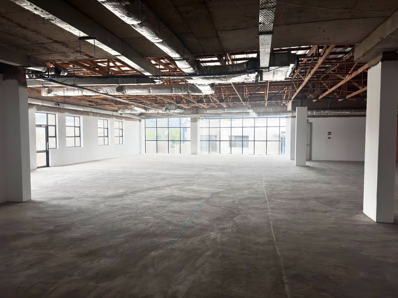 A spacious, open-plan office area featuring an industrial aesthetic with exposed ceilings, ductwork, and large windows providing ample natural light.