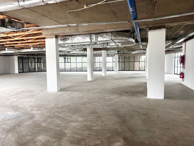 A large, open-plan office space in shell condition featuring exposed concrete ceilings, structural columns, and expansive glass windows.
