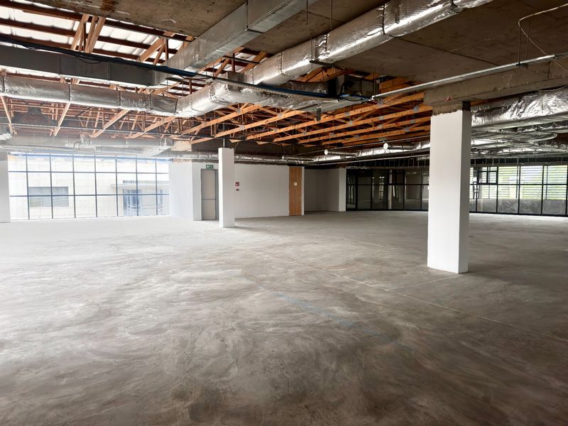 A large, open-plan office space in shell condition, featuring exposed industrial ceilings, concrete flooring, and expansive windows providing ample natural light.