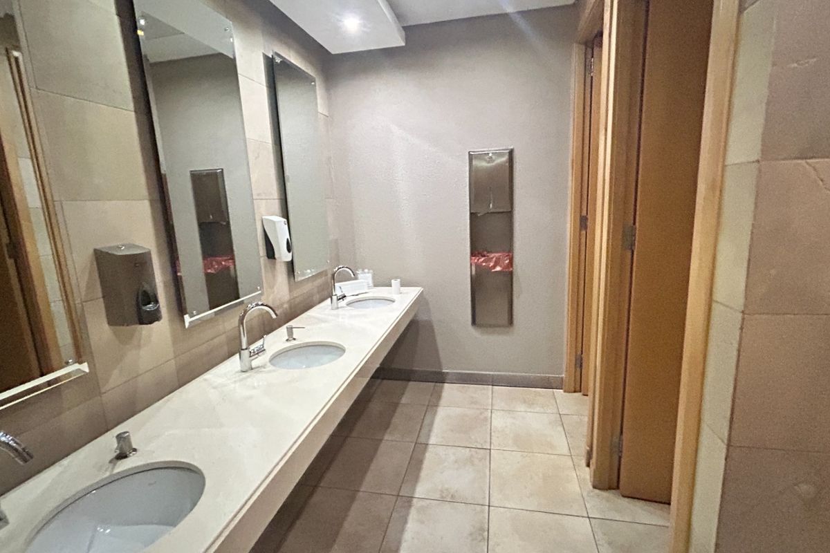 A clean, modern office bathroom featuring a long vanity with multiple undermount sinks, chrome fixtures, tiled surfaces, and private cubicle stalls.