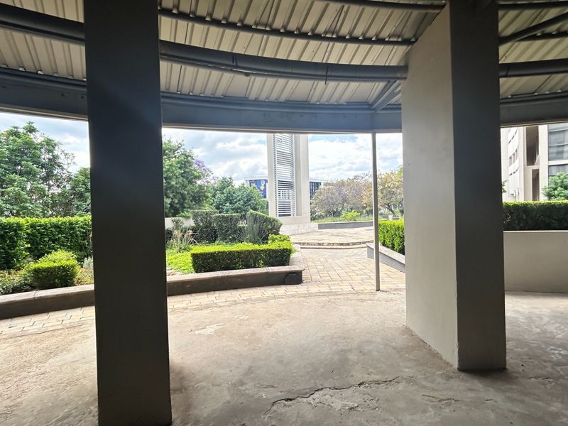 A covered outdoor patio area featuring concrete pillars and a corrugated metal roof, overlooking a landscaped garden and paved walkway.