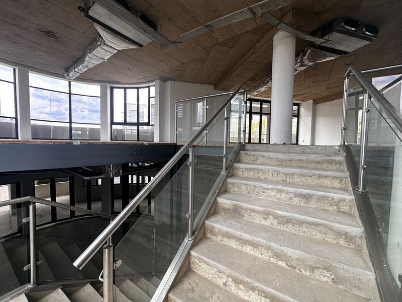 An unfinished open-plan office space featuring a concrete staircase with glass balustrades, exposed ceiling ductwork, and large windows providing ample natural light.