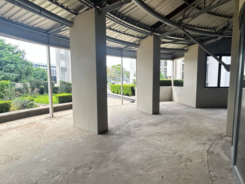 An expansive, unfinished outdoor patio area featuring exposed steel roofing, concrete support columns, and views of the surrounding landscaped garden.