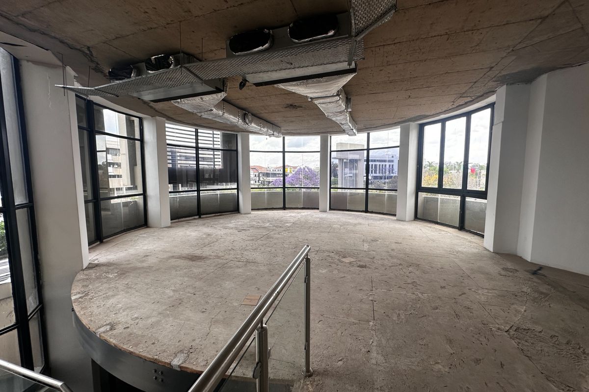 An expansive, unfinished office space featuring a curved layout, floor-to-ceiling windows with city views, and exposed industrial ceiling ductwork.