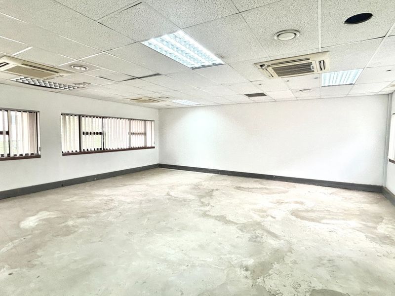 An empty, open-plan office space featuring a suspended ceiling with integrated lighting, cassette air conditioning units, and large windows with vertical blinds.