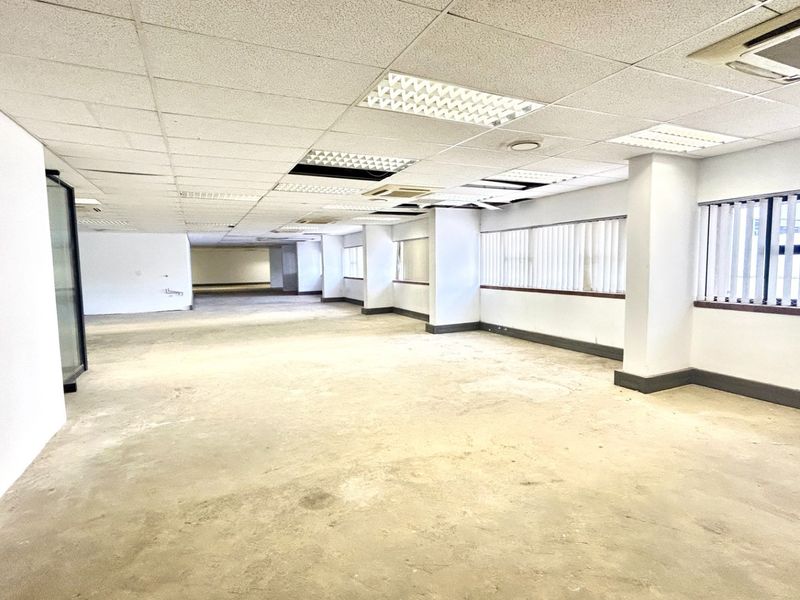 A large, open-plan office space featuring a suspended ceiling, recessed lighting, and vertical blinds, currently in need of flooring and ceiling repairs.