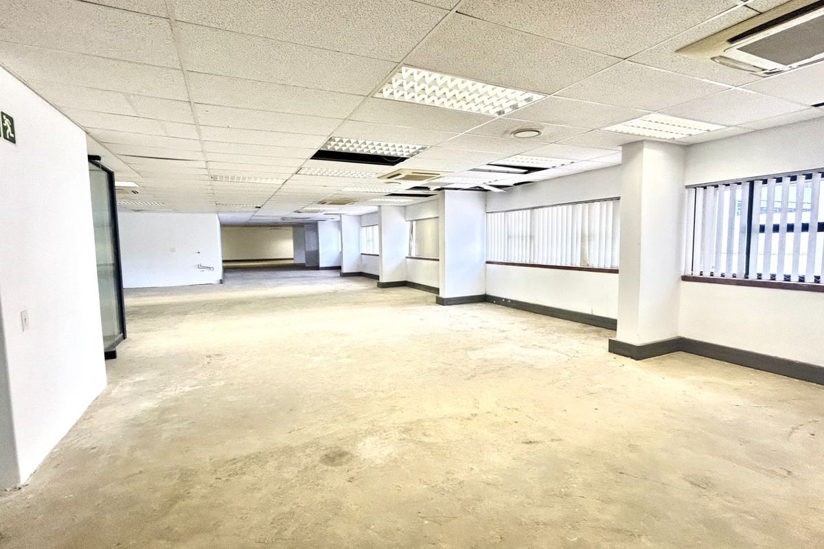 A large, open-plan office space featuring a suspended ceiling, recessed lighting, and vertical blinds, currently in need of flooring and ceiling repairs.