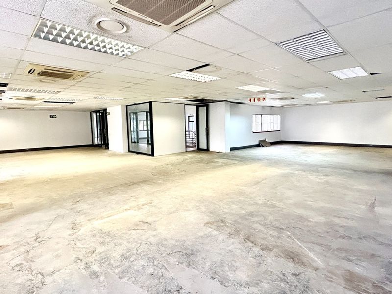 A large, open-plan office space featuring a suspended ceiling with recessed lighting, air conditioning units, and glass partitions, currently in need of flooring refurbishment.