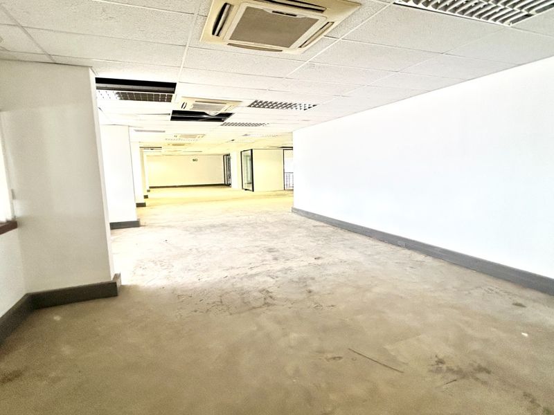 A large, vacant open-plan office space featuring a suspended ceiling with integrated air conditioning and large windows with vertical blinds.