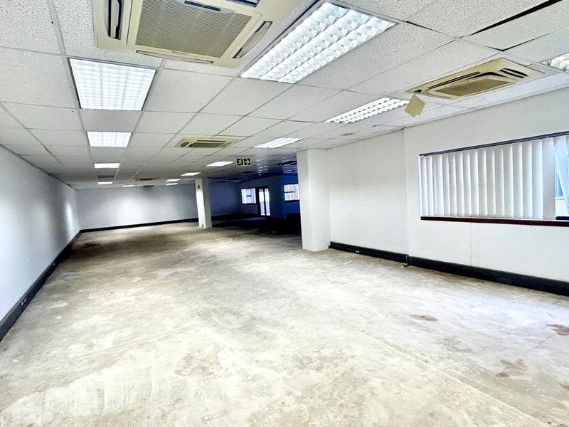 An expansive, vacant open-plan office space featuring a suspended ceiling, integrated air conditioning, and large windows with vertical blinds.
