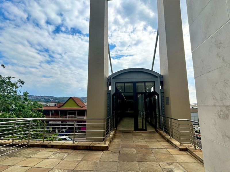 An elevated stone-paved terrace featuring a modern glass and steel walkway structure with stainless steel balustrades, offering an open outdoor connection.