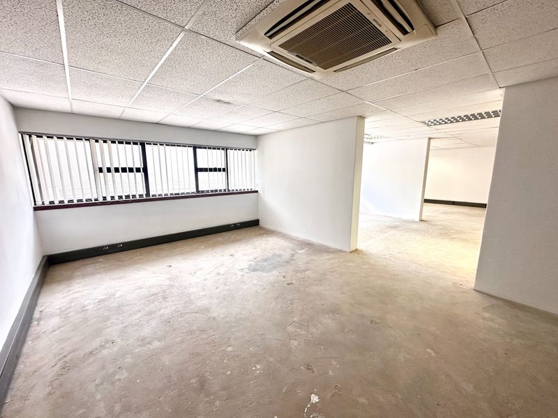 An empty, open-plan office space featuring a suspended ceiling with integrated air conditioning, vertical blinds, and unfinished concrete flooring.